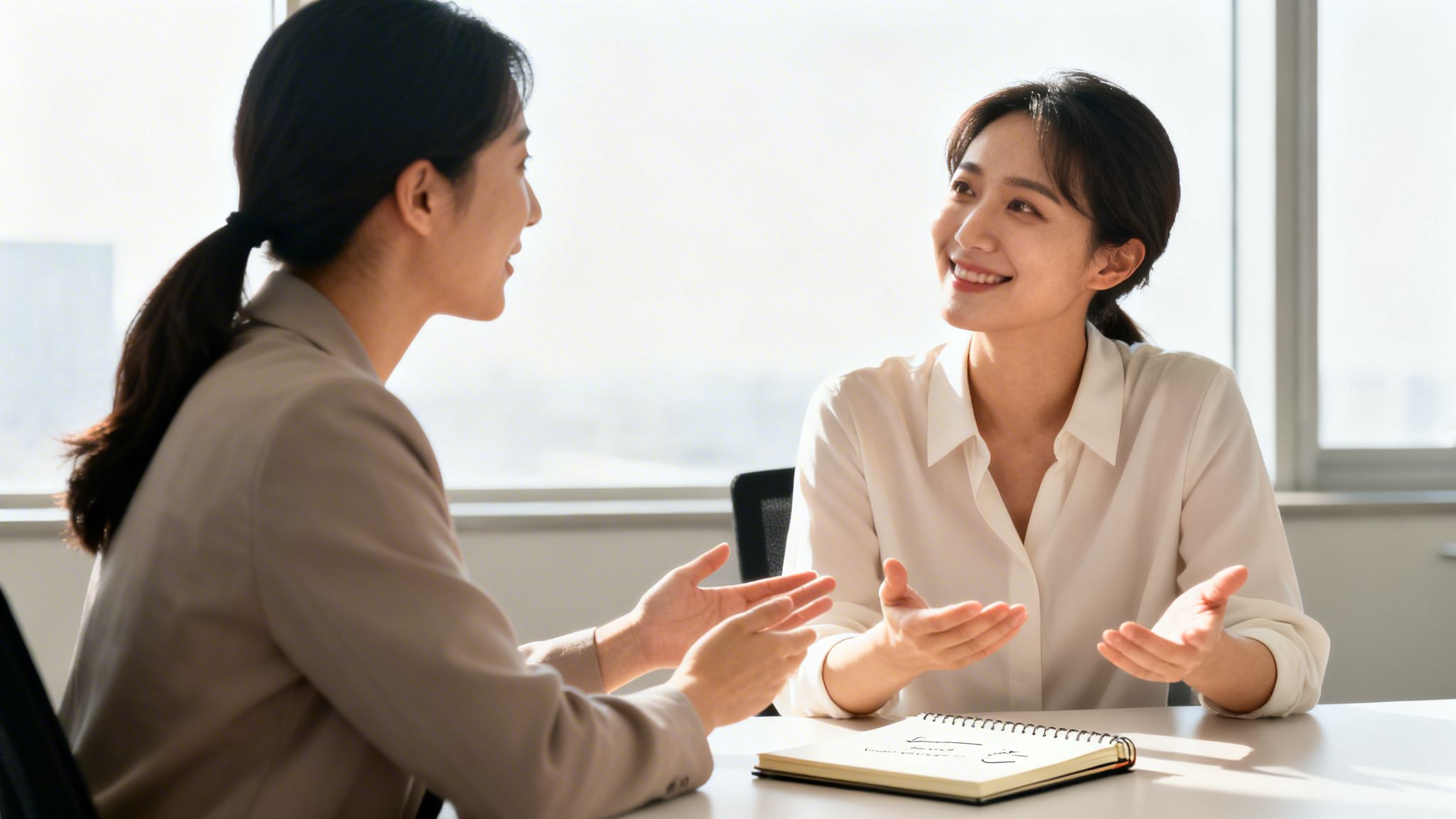 Smiling professional women discussing ideas in a sunlit modern office with a notebook.