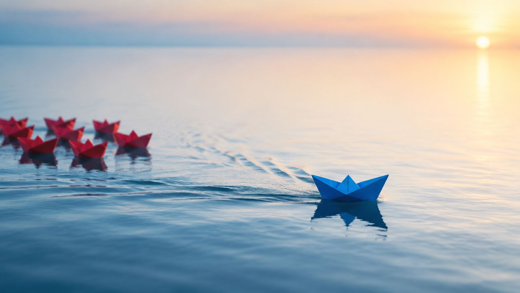 A blue paper boat leading a group of red paper boats on calm water at sunrise.