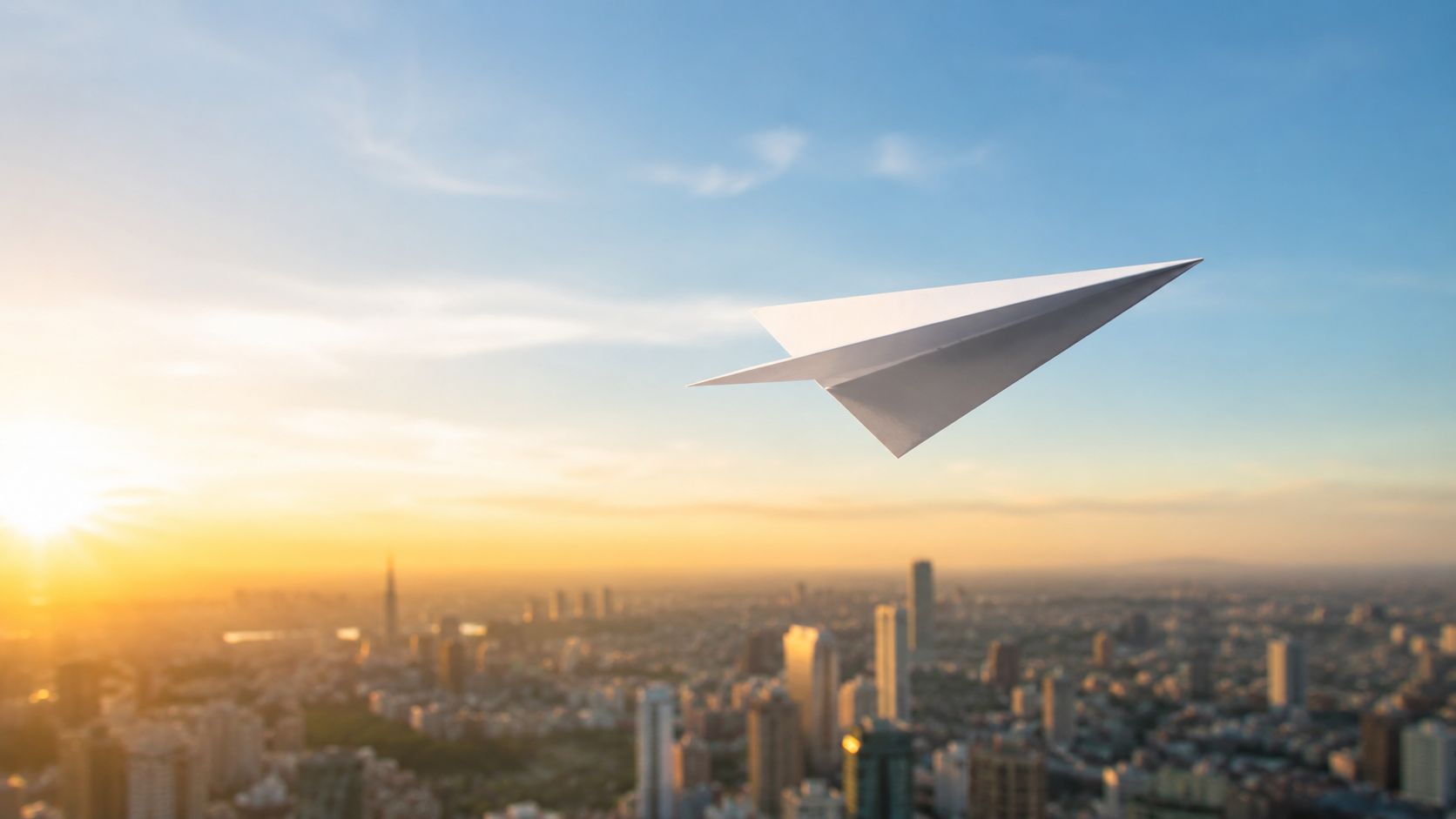 A paper airplane flying over a large city skyline during a beautiful orange sunset at dusk.