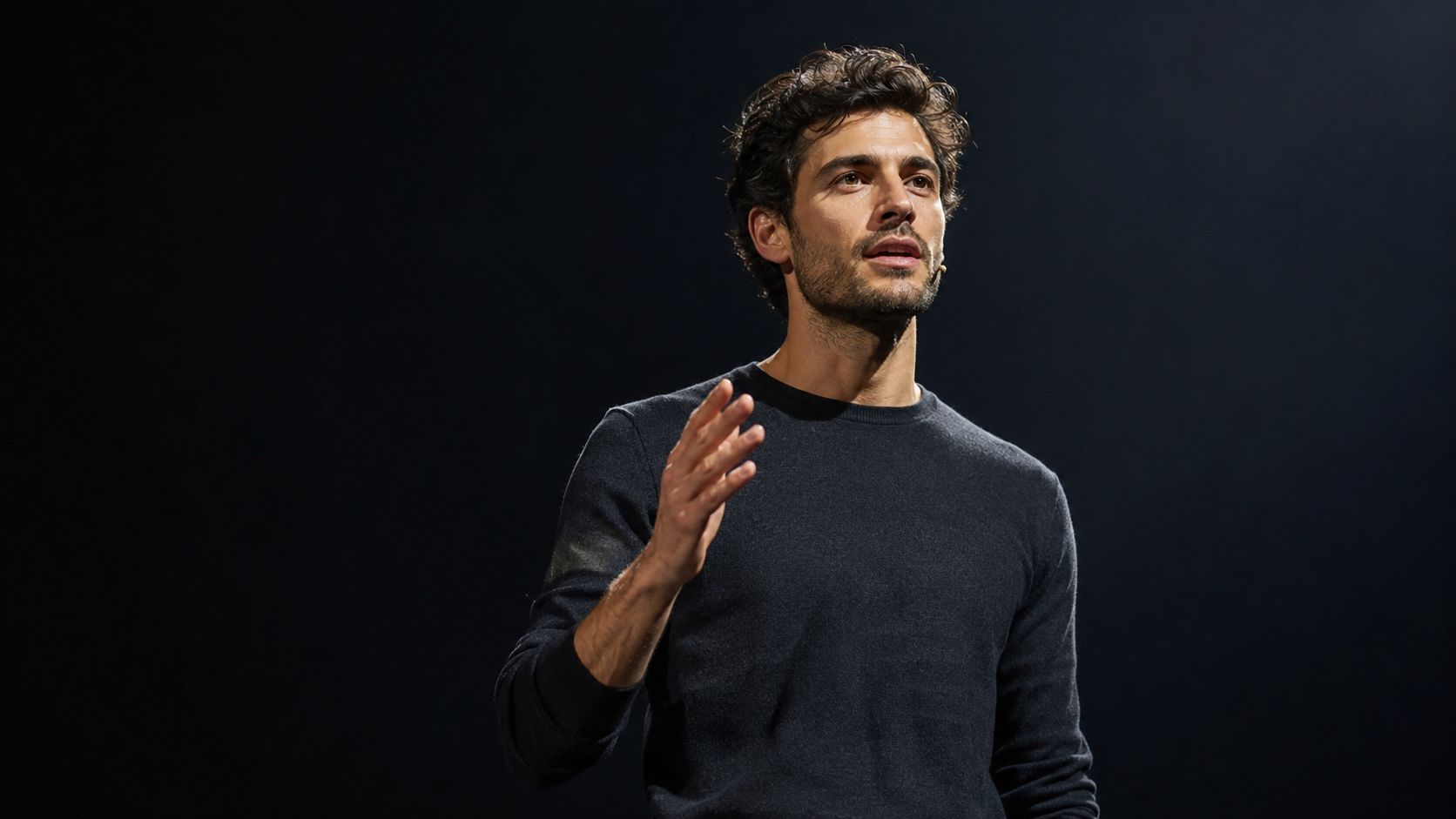 A young man with wavy dark hair and a beard speaking while wearing a dark grey sweater.